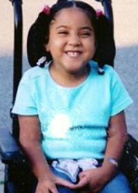 Photo of Julie Cirella, a smiling girl wearing pigtails and sitting in a power chair. She has dark skin, and is wearing a teal T-shirt.
