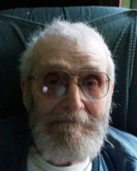 Photo: An older man with a full gray beard and hair; he is fair-skinned, balding, wearing tinted glasses.