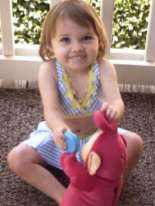 Photo of Katie McCarron, a toddler girl with blond hair and pale skin, wearing a blue summer dresss, sitting on the carpet and playing with a Teletubby toy.