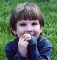 Photo of Marcus Fiesel, a small boy on a green lawn. He has pale skin and windswept brown hair in a bowl cut. He is shyly covering his mouth with his hands.
