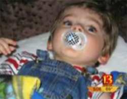Photo of Payton Ettinger, a toddler boy lying on a mat and sucking on a pacifier; he has pale skin and blond hair, and is wearing denim overalls and a striped shirt.