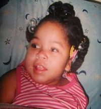 Photo of Shylae Thomas, a girl with her hair in braids and barrettes, propped up against a cushion. She has light-brown skin and dark-brown hair, and her shirt is a red and white striped tank top.