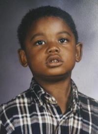 "School photo of a young boy with brown skin and short, curly black hair, wearing a black and white checked shirt.