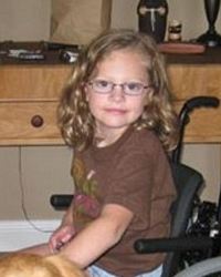 Photo of a young girl sitting in a wheelchair; she has curly blond hair, fair skin, and glasses, and she is wearing a brown T-shirt.