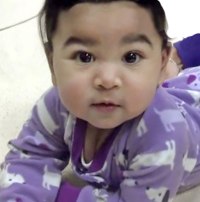 Photo: An Asian toddler lying on her front, propped up on her arms. She is wearing a purple printed onesie.