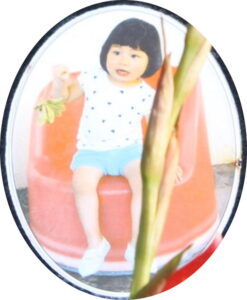 Photo of a girl with short, straight black hair and fair skin, sitting in a red plastic chair; she is wearing a polka-dirt shirt and blue shorts.