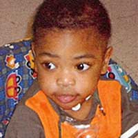 Photo of a toddler in  a baby seat. He has large brown eyes, brown skin, and wispy black hair. He is wearing an orange shirt.