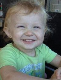 Photo of a toddler with wispy blond hair and fair skin, wearing a green shirt, grinning at the camera.