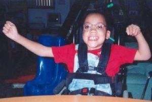 Photo of Danieal Kelly, a girl sitting in a wheelchair, smiling and waving her arms. She has dark hair and light-brown skin, and is wearing a red T-shirt with denim overalls.
