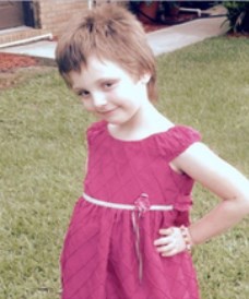 Photo of a little pale-skinned girl in a pink dress. Her brown hair is cut short in a boy cut. She has her hands on her hips and is smiling slightly.