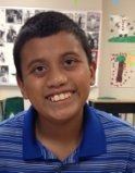 Photo of a young boy with brown hair and tan skin, smiling; in the background are the walls of a school classroom.