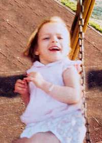 Photo of Andrea Lago, a blonde toddler sitting on a swing. She is wearing a white shirt and short pants, and smiling.