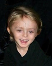 Photo of a toddler with short, light hair against a black background. His head is larger than average. He is smiling.
