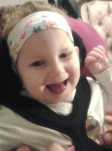 Photo of a baby girl with curly blond hair tied in a headband, strapped into a car seat. She is smiling.