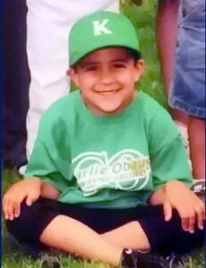 Photo of a boy in a green shirt and baseball cap; he has tan skin, and is smiling, sitting cross-legged on the ground.
