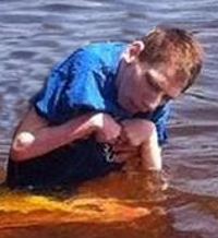 Photo of Michael Furst. He is a young man being held up by someone behind him; he is up to his waist in water and leaning over to look into the water. He has red hair and fair skin; his arms are curled over his chest because of spasticity. He wears a blue T-shirt.