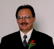 Photo of Ronald McCabe. He is a middle-aged man wearing glasses and a suit with a rose pinned to the lapel. He has fair skin, dark brown hair and a mustache.