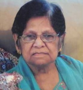 Photo of an older woman in a turqoise top; she has olive skin and black hair with gray roots. She is wearing glasses.