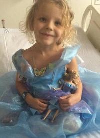 Photo of Emily Perrin, a small blonde girl sitting on a bed. She is wearing a light blue dress and holding a doll wearing a matching blue dress.