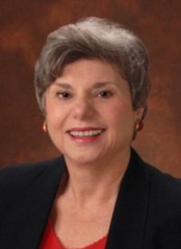 Portrait photo of Marie Stempinski. She is a woman with pale skin and gray hair, wearing business attire and chunky red earrings.