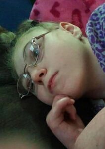 Photo of a teenage girl with pale skin and dark-blond hair in a scruffy ponytail; she is wearing glasses, lying down, her chin in her hand.