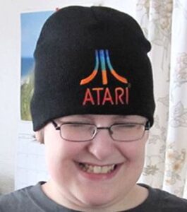 Photo of a fair-skinned teenage boy, smiling; he is wearing square-rimmed glasses and a black beanie with the Atari logo.