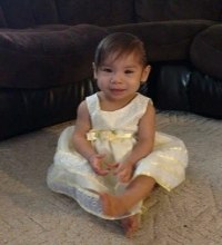 Photo of Kira Friedman. She is a toddler with straight brown hair and tan skin. She is barefoot, wearing a fancy white dress.