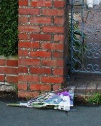 Flowers left at Jean Irwin's house. They are purple and white flowers, lying on a curb next to a brick wall and wrought-iron gate.