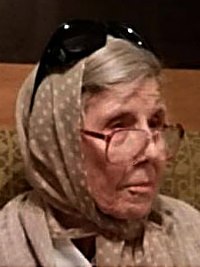 Photo of Peggy Sinclair. She is an elderly woman with light skin and gray hair; she is wearing glasses, which are slipping down her nose, and a polka-dotted beige headscarf.