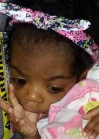 Photo of Jazmine Walker, a small infant with dark skin and curly black hair. She is wearing a pink headband and dress, and is sucking her thumb.