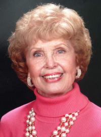 Photo of Nada Bodholdt. She is an elderly woman with strawberry blonde curly hair, cut short. She is wearing a pink turtleneck, earrings and a necklace.