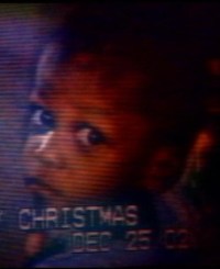 Photo of Deetrick Brown. It is a photo of a TV screen, showing an African-American toddler with a solemn expression. Faint text along the bottom says, "Christmas. December 25." The rest of the text is indistinct.