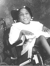 Black and white photo of Janelle Johnson. She is a young African-American girl in a white dress, sitting in a power wheelchair and smiling.