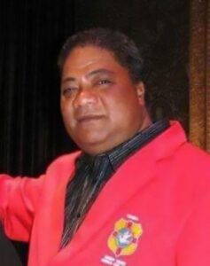 Photo of a man in a bright red suit jacket and black shirt and tie. He has brown skin and short black hair; his nose is broad, and his face is round.