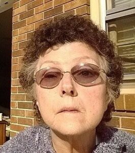 Photo: An elderly woman with curly brown short hair and pale skin, photographed against the brick wall of a house; she is wearing sunglasses. Her expression is neutral. 