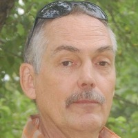 Photo of John Owings against a background of leaves. He is a fair-skinned older man with graying hair and mustache. He has sunglasses propped up on his head.