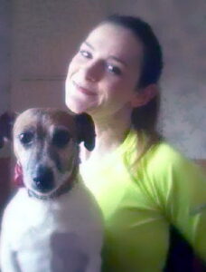 Photo: A young woman in a bright yellow T-shirt, her brown hair in a ponytail. She has pale skin; her head is tilted. In the foreground is a small brown and white terrier dog.