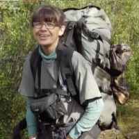 Photo of Riba DeWilde. She is a woman with brown hair, fair skin, and large glasses. She is wearing a large hiking backpack and smiling.