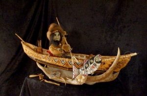 Photo of "Athabaskan Male Doll with Accessories", artwork by Riba Dewilde. The doll is made of wood, bark, fur, and antler. It is dressed in traditional clothing, sitting in a canoe and holding a fishing spear.