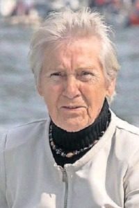 Photo of Rietje Willms, an elder woman. She has short white hair and fair skin, and is wearing a black turtleneck and white jacket. Her expression is solemn, and she is squinting slightly into the sun.