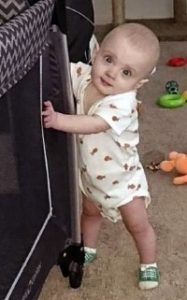 Photo of Aedyn Agminalis, a baby with fair skin and wispy blond hair. He is wearing a onesie and holding on to a piece of furniture to support him as he stands on wobbly legs.