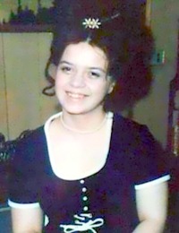 Photo of Perleen Bode. She is a young woman with fair skin and dark curly hair; she is wearing an old-fashioned navy blue dress with white trim.