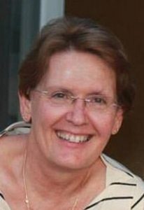 Photo of Pamela Kruspe, an older woman with fair skin and brown hair. She is wearing glasses and a striped shirt; she is smiling at the camera.