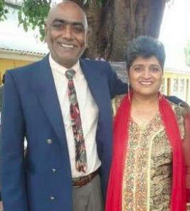 Photo of Prakash and Shoba Singh. He is a middle-aged man wearing a suit and tie; his skin is medium-brown and his head is shaved. She is wearing a dress with a red sash, has light-brown skin and curly short gray hair.