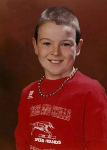 Photo of Ian Carmichael, a boy with fair skin and short brown hair. He is wearing a red sweater with a sports logo on it, and a necklace of steel-gray beads. He is smiling for the camera.