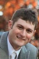 Photo of Mark Mellor, a young man wearing a gray suit and tie. He has fair skin with ruddy cheeks and short brown hair; he is smiling at the camera.