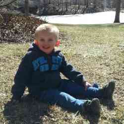 Photo of Brayson Price, a young boy with short-cropped blond hair and fair skin, wearing jeans and a blue jacket. He is sitting outside on a grassy lawn.