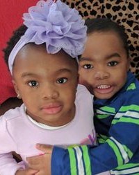 Photo of Olivia and Micah Gee, two African-American toddlers. Olivia is wearing a pink sweater and has her hair tied back in a headband with a big purple flower on it. Micah is wearing a striped blue-and-green shirt, and has his arms around his sister.