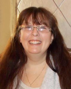 Photo of Donita Elliott, a middle-aged woman with fair skin and very long dark-brown hair, wearing glasses, a white sweater, and a gold necklace. She is smiling broadly at the camera.