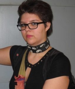 Photo of Ryanna Grywacheski, a young woman wearing dark-rimmed glasses, a choker, and a dark blouse. Her hair is dark brown and cut short; her skin is fair. She is wearing light makeup.